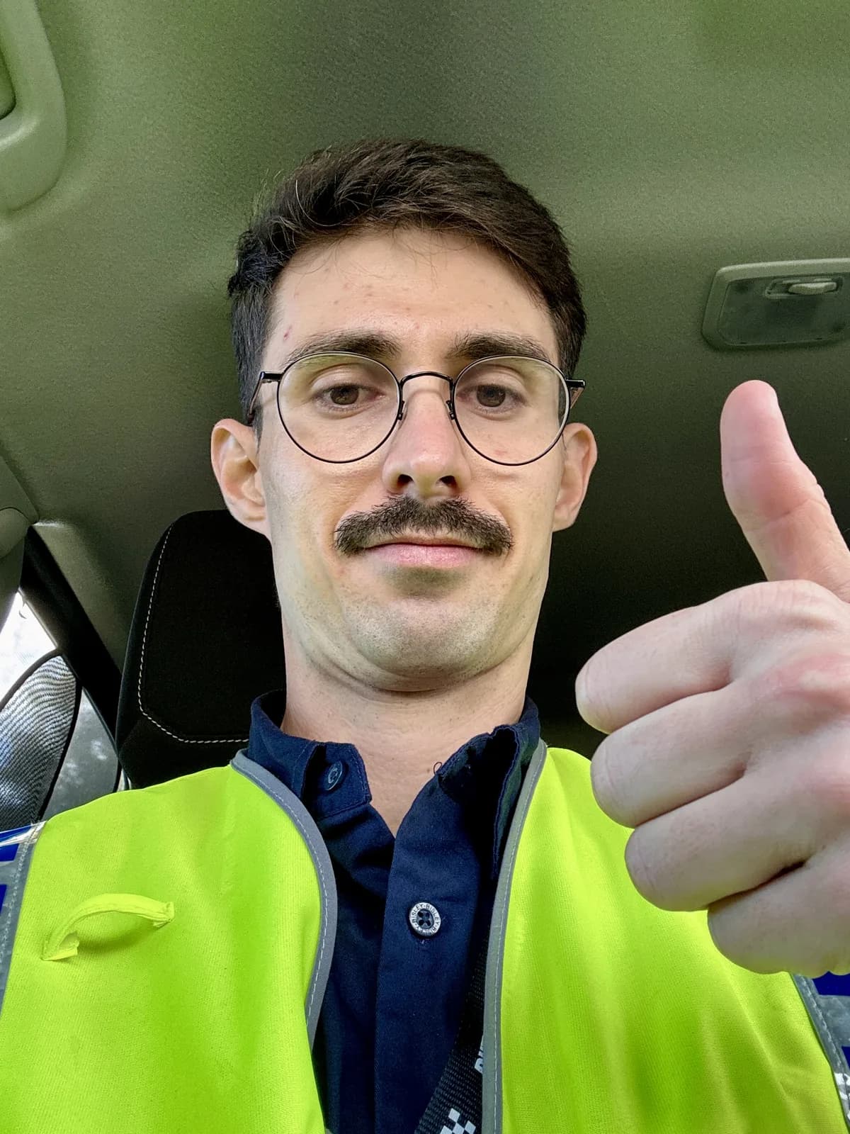Lennox in a hi-vis vest in the driver’s seat of a car, giving a tired thumbs up